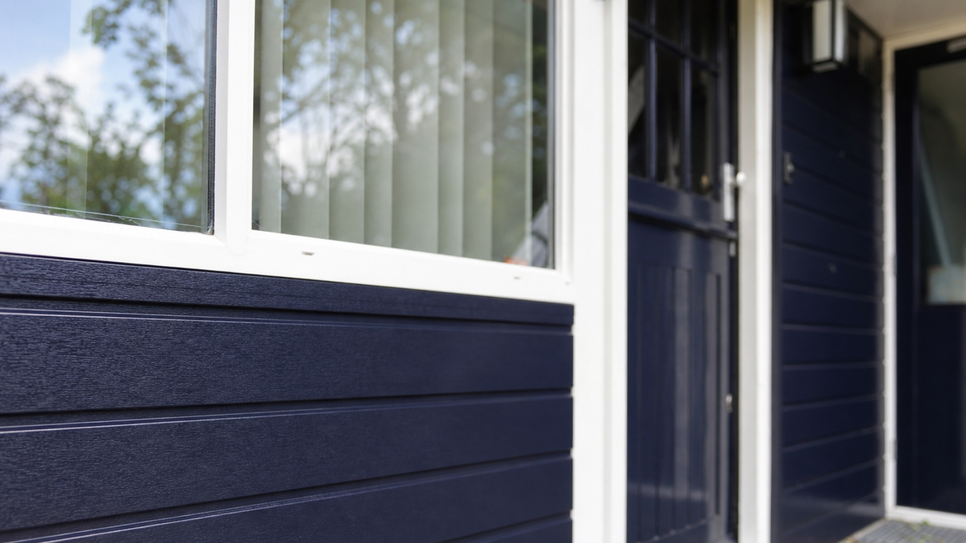 Staalblauwe kunststof gevelbekleding onder een wit kozijn bij de entree van een woning.